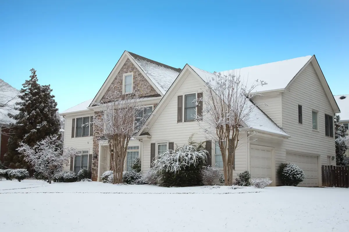 A two-story suburban house with siding insulation, its snow-covered roof and yard surrounded by trees and bushes, under a clear blue sky.
