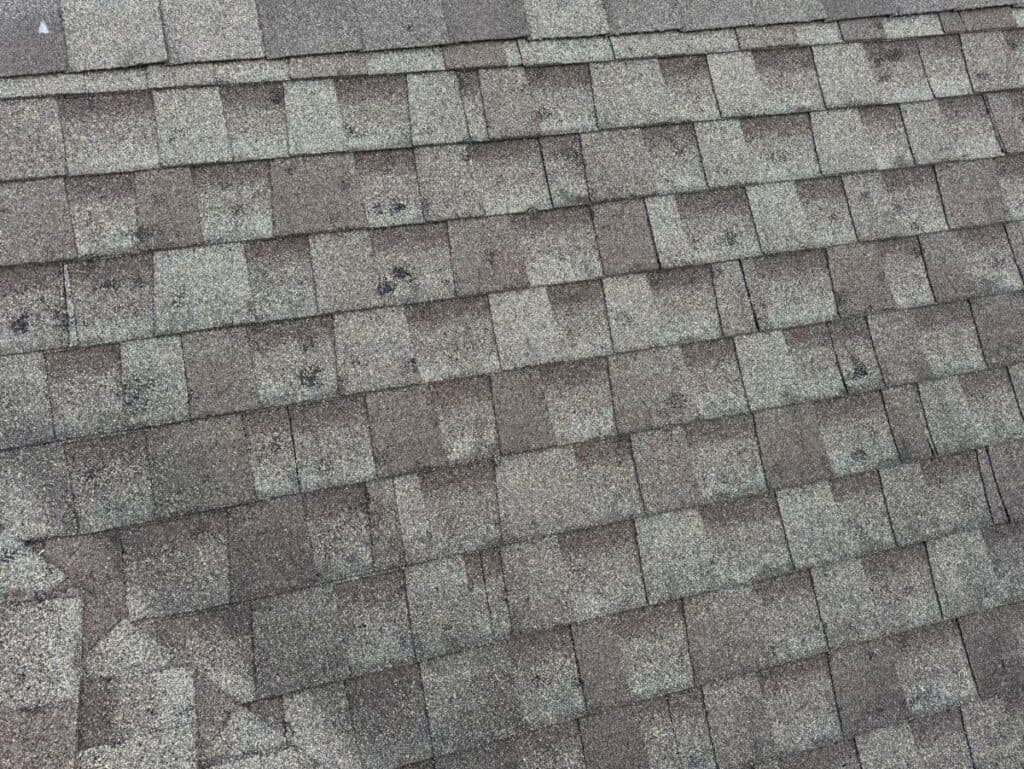 storm damage roof repair Close-up view of asphalt roof shingles in alternating light and dark gray tones, showing a pattern of overlapping rectangular shapes with some visible dark spots and wear.