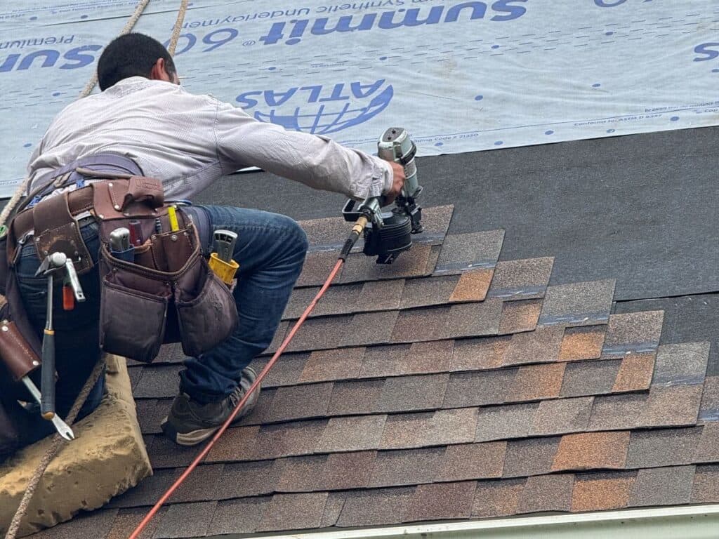 storm damage roof repair Two workers install shingles and underlayment on a sloped roof. One is using a nail gun, wearing a tool belt, while the other sits higher up, partially visible. The roof is partially covered with synthetic underlayment.