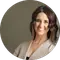 A person with shoulder-length hair is smiling, wearing a light-colored top. The background is a plain, muted color. The image is circular.