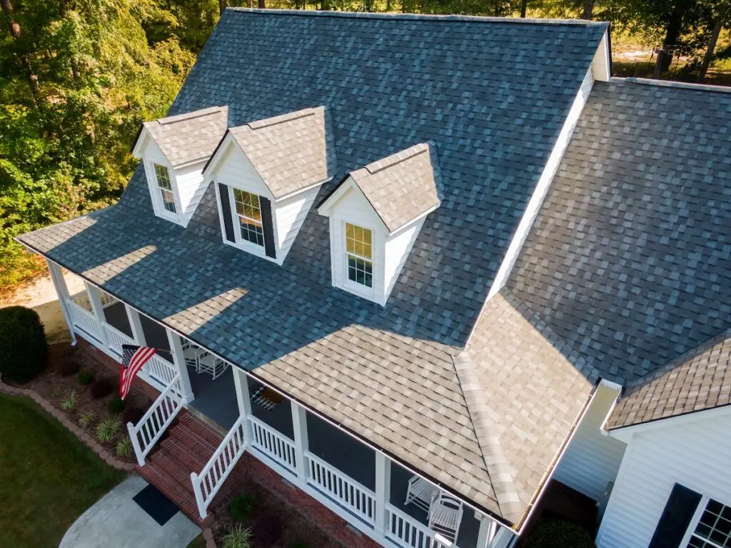 Aerial view of a house with gray shingle roofing, white siding, and three dormer windows—an ideal example when considering metal roof vs shingles. A front porch with a white railing and an American flag is surrounded by green trees and landscaping. metal roof vs shingles