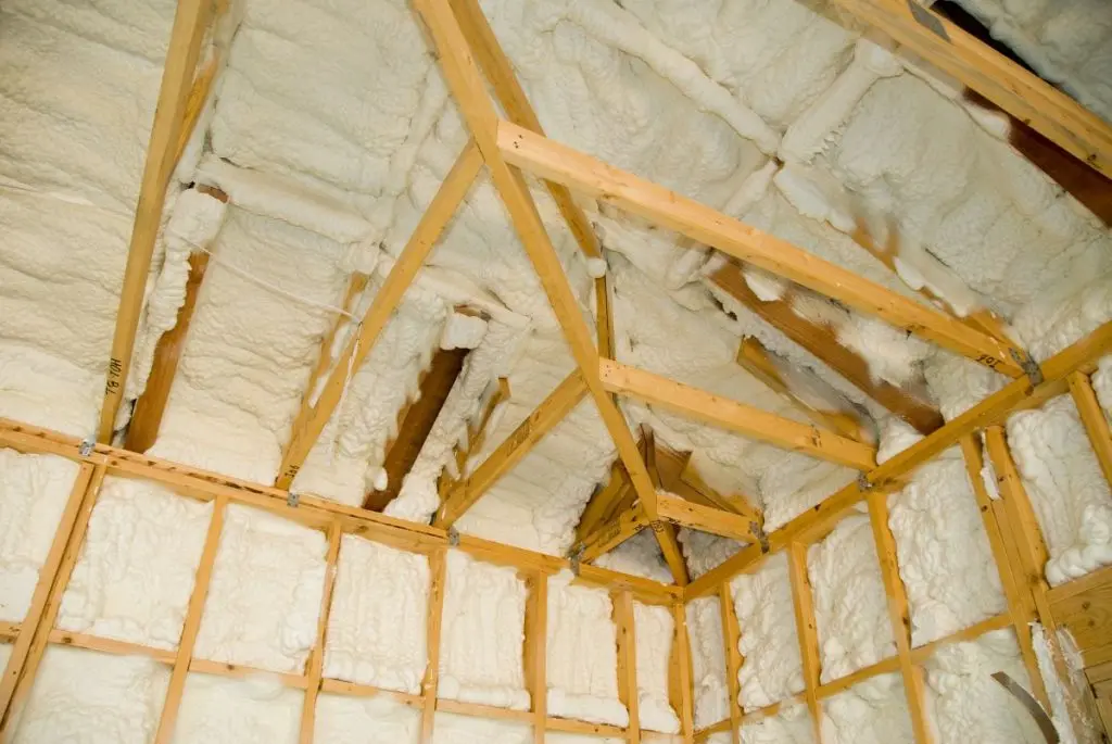 Wooden framing of a house under construction with white spray foam insulation applied between the beams on walls and ceiling.