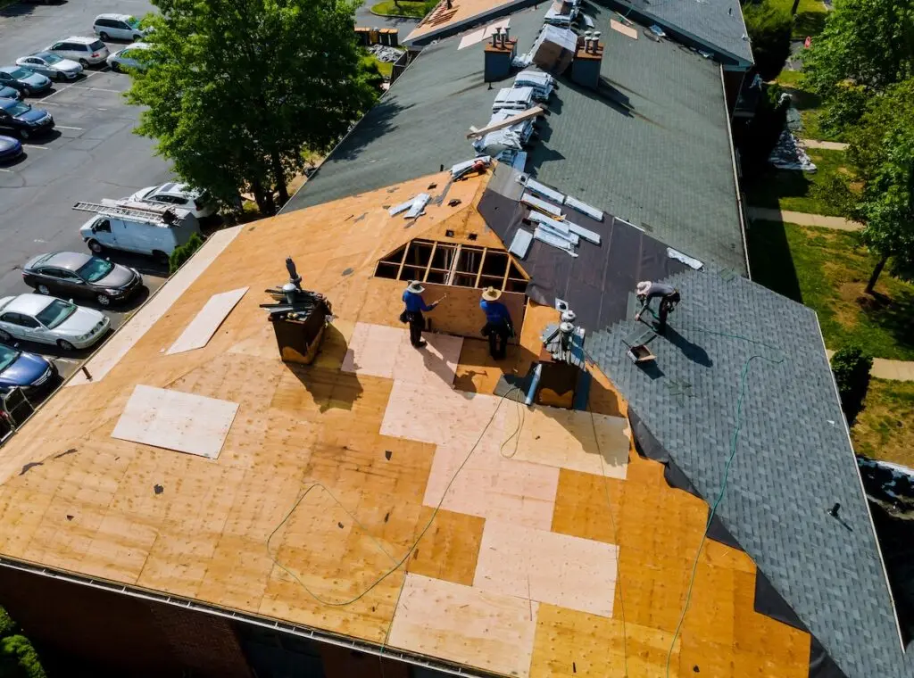 Aerial view of a building roof under repair. Three workers are installing plywood sheets, with green shingles partially in place. Construction equipment and materials are scattered across the roof. Parked cars and trees are visible on the left.