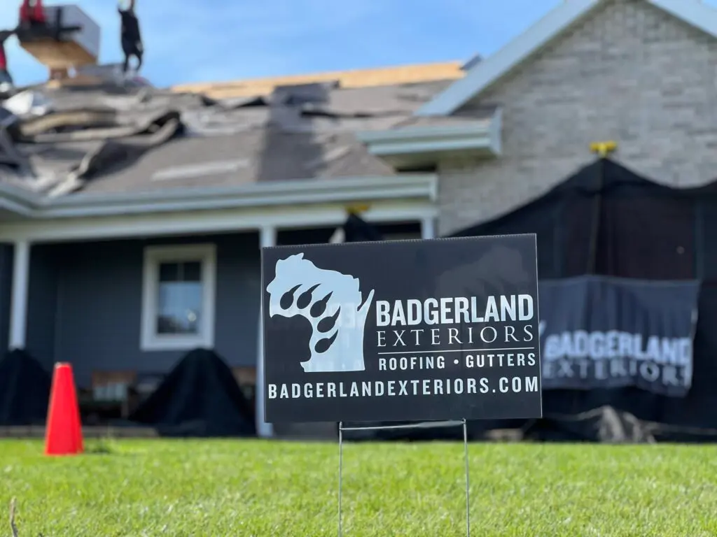 A Badgerland Exteriors sign is displayed in a yard, with workers on the roof of a house in the background. The house is under construction with tarps and tools visible.