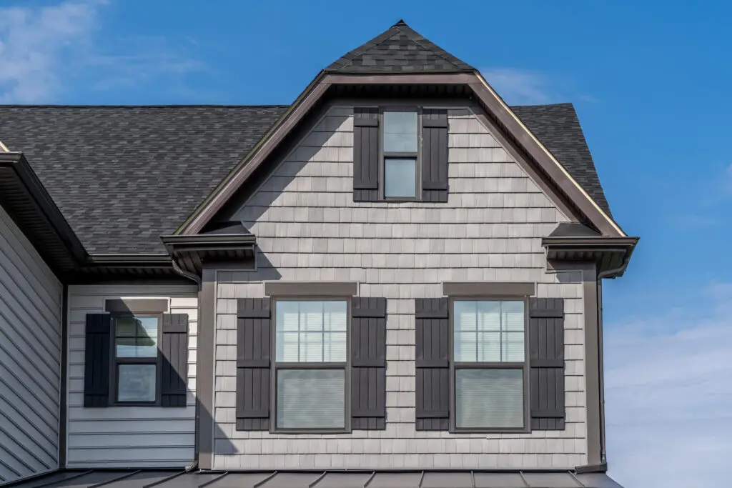 The image shows the exterior of a house with white siding and black shutters. It features two windows on the upper story and a steep, dark-colored roof against a clear blue sky.