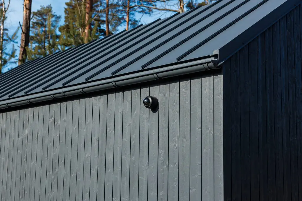 A modern building with dark vertical wooden siding, a metal roof, and a security camera mounted on the wall. Tall pine trees and blue sky are visible in the background.
