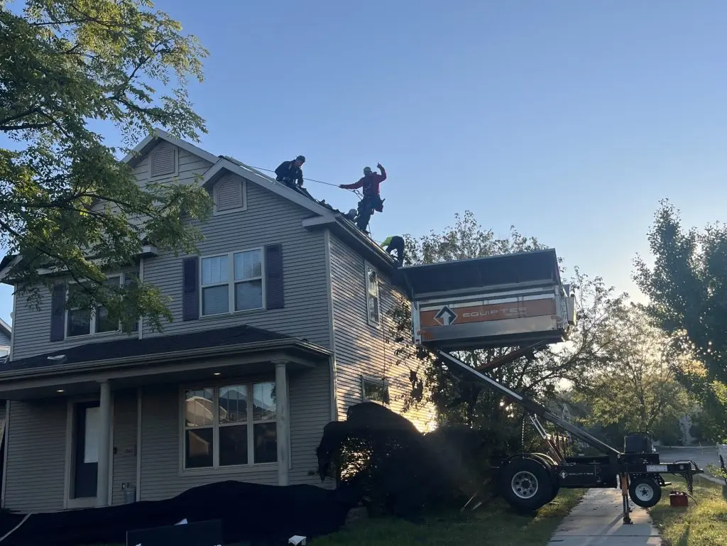 Two workers repair the roof of a two-story house at sunrise, with equipment and a trailer parked outside. A sign in the yard reads “Badgerland Exteriors.” Trees and a clear sky are in the background.