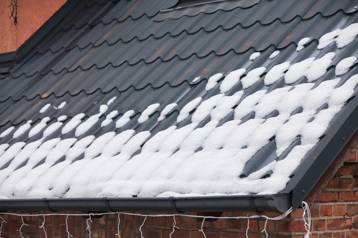 A sloped black metal roof, expertly maintained with metal roof leak repair, is partially covered with patches of snow above a brick wall. White holiday lights hang from the edge of the roof.