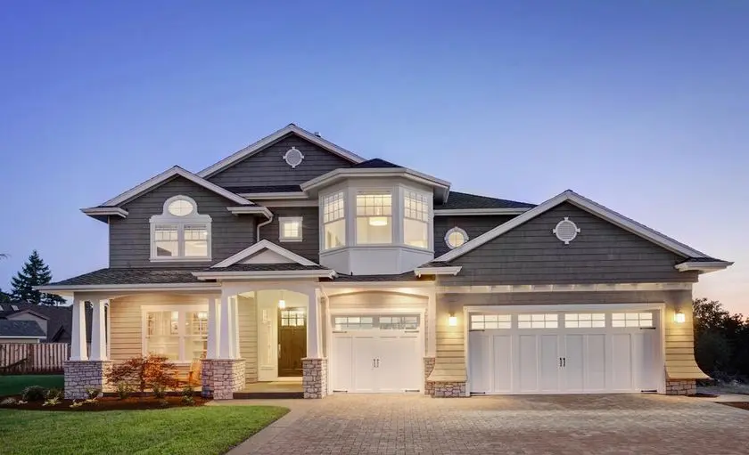 A large, modern two-story house in Madison showcases a gray exterior with white trim and expertly crafted roofing. It features a symmetrical design with two garage doors, expansive windows, and a well-lit entrance against the backdrop of a clear early evening sky.