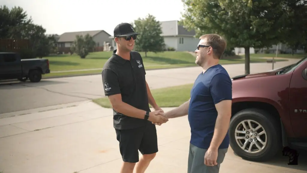 Two men shake hands and smile while standing in a driveway next to a red SUV on a sunny day, with houses and green lawns in the background.