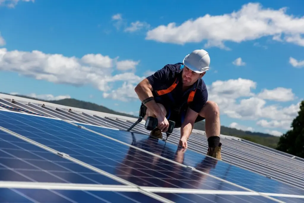 A worker wearing a white hard hat and safety harness installs solar panels on a rooftop in Verona under a blue sky with a few clouds. The scene conveys a focus on renewable energy and technology, highlighting the rise of solar installation in this picturesque Italian city.