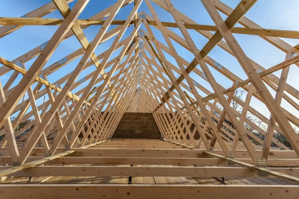 Wooden roof trusses form a triangular pattern against a blue sky. The skeletal framework of a building under construction is visible, with beams connecting at the peak and extending down to the floor level. Sunlight casts shadows across the structure.