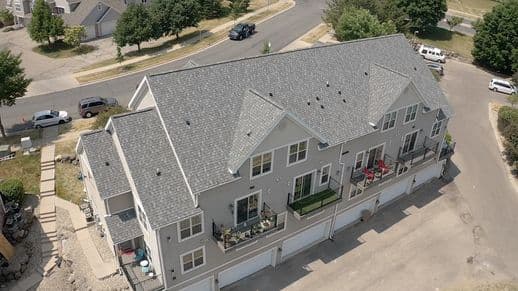 An aerial view of a large, two-story residential building with a gray roof. The building features multiple balconies and garages. Surrounding streets and parked cars are visible, along with some greenery in the neighborhood.