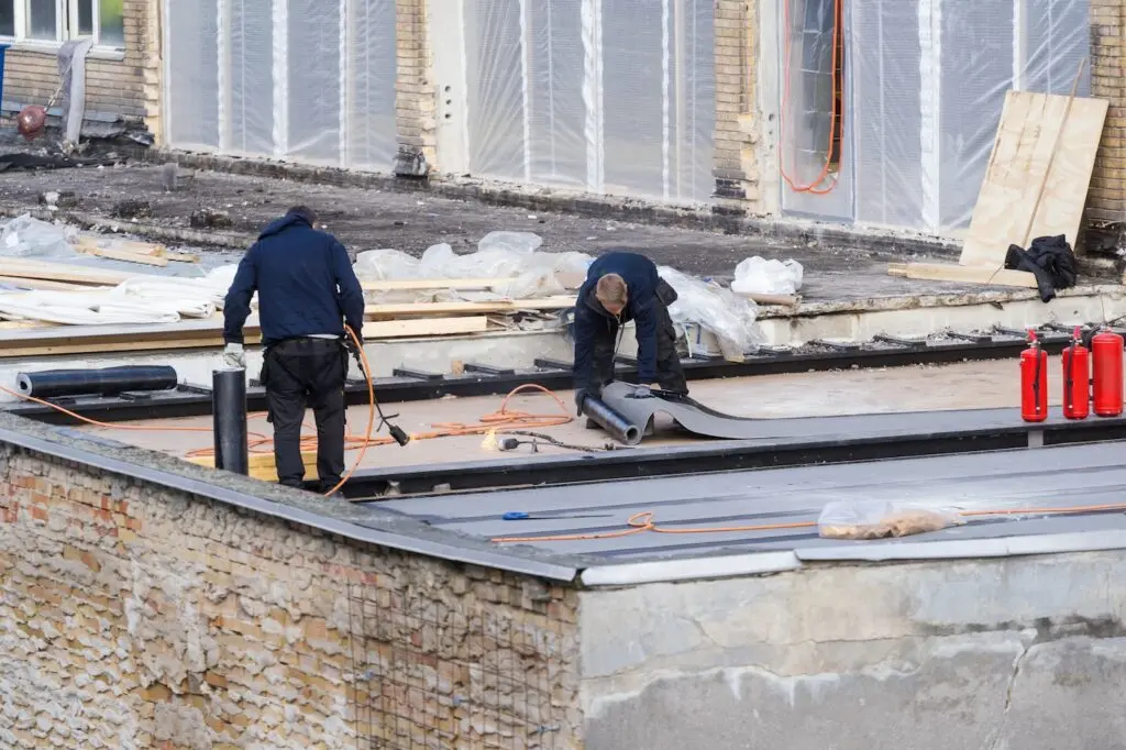Two workers on a rooftop install or repair roofing material, using tools and black roll material. The roof is surrounded by construction supplies, fire extinguishers, and building walls covered in plastic sheets.