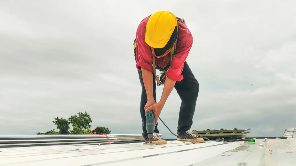 types of commercial roofing systems A worker wearing a yellow hard hat and red shirt uses a power drill on a metal roof under a cloudy sky. Trees are visible in the background.