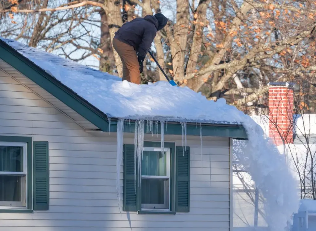 A person in winter clothes shovels snow from the roof of a house with green shutters. Long icicles hang from the roof’s edge, and bare trees are visible in the background.