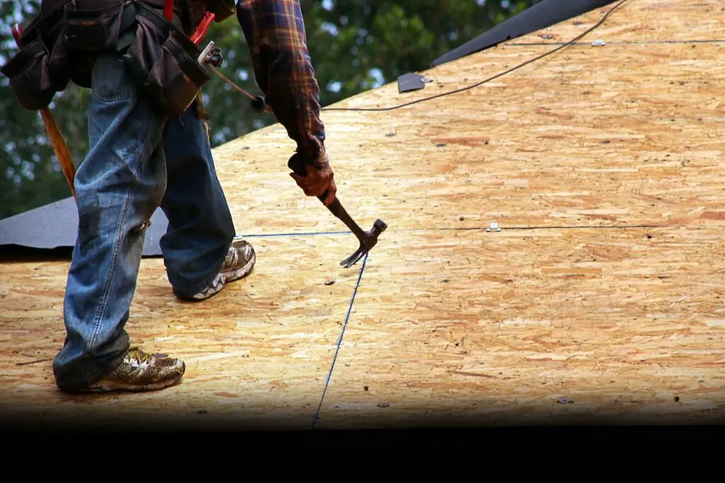 A construction worker in jeans and a plaid shirt uses a hammer on a plywood roof. The worker wears a tool belt and sneakers, and is visible from the waist down. The roof is partially covered with black felt paper.