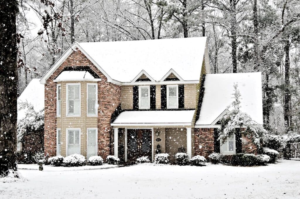 siding insulation A two-story house with brick and beige siding insulation is surrounded by trees and covered in snow. Snow is falling, blanketing the roof, yard, and shrubs, creating a peaceful winter scene.