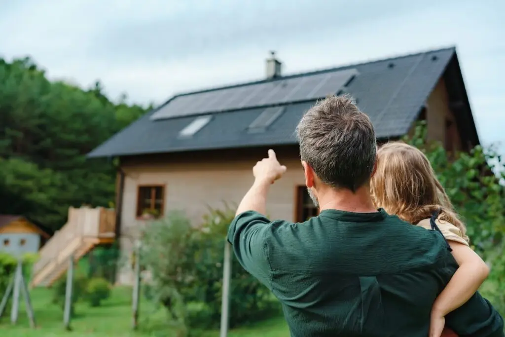 A man holding a child points towards solar panels on the roof of a wooden house surrounded by greenery. The scene is bright and outdoors, echoing an eco-friendly lifestyle that rivals the best of commercial roofing Madison has to offer in sustainable solutions.