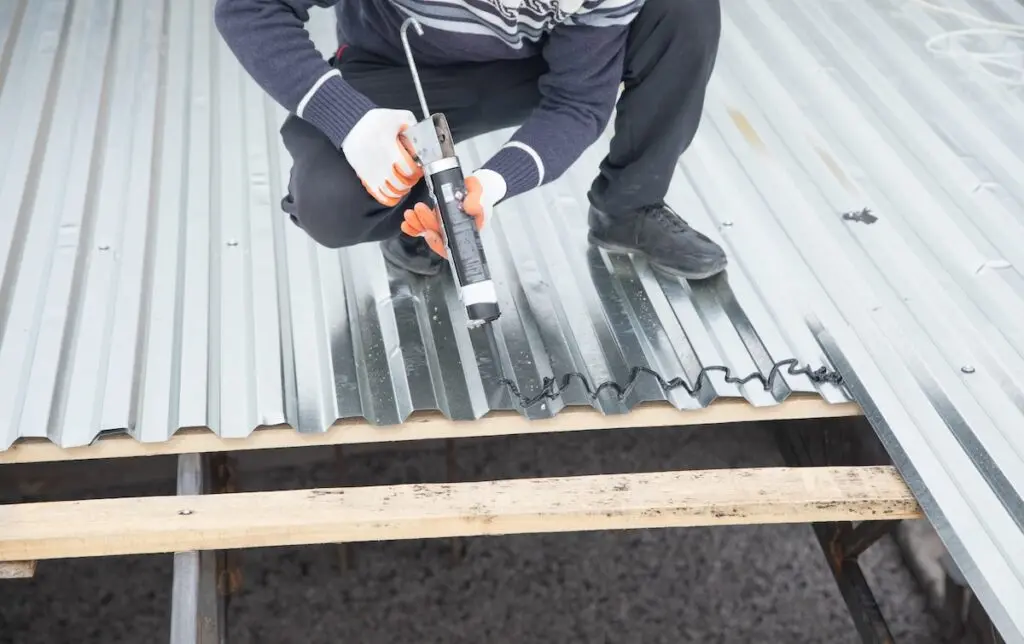 waterproof sealant for metal roof A person wearing gloves kneels on wooden beams, carefully applying waterproof sealant for metal roof to the seam of a corrugated metal roof panel.