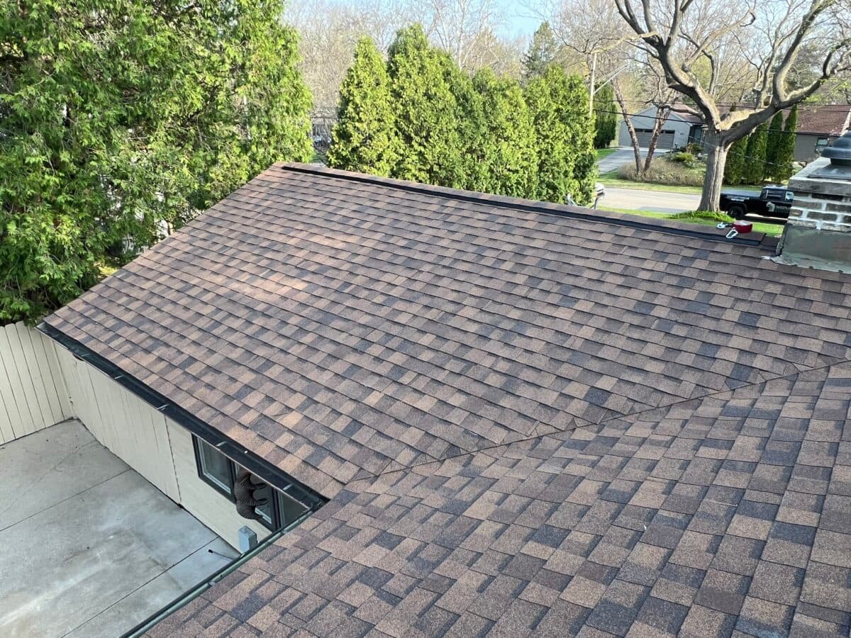 asphalt shingle roof replacement A house roof with brown asphalt shingles, seen from above. Surrounding the roof are green trees and part of a paved driveway. The scene appears to be in a residential neighborhood.