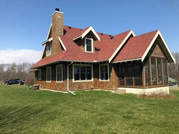 A two-story house with a red roof and wooden siding, featuring a chimney and large windows. It has a screened porch on the right side and is set on a grassy lawn under a clear blue sky. A car is parked nearby.