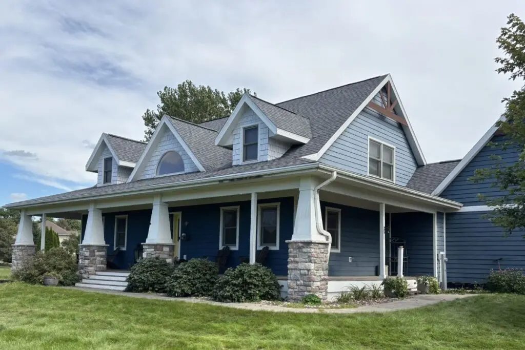 Blue two-story house with white trim, stone pillars, a large front porch, and multiple gable roofs showcases expert roofing Sauk City craftsmanship. Green shrubs line the front on a well-kept lawn under a partly cloudy sky.