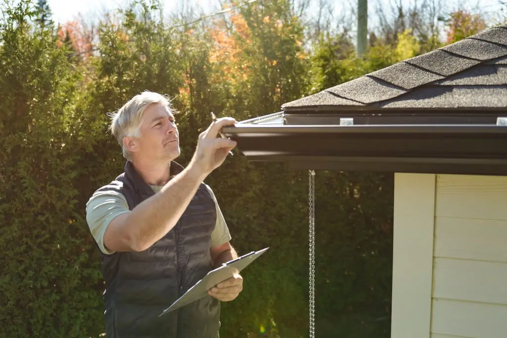 how to prepare your roof for winter A man holding a clipboard inspects the roof and gutter of a house on a sunny day, with trees and shrubbery in the background.