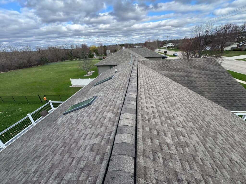 roofing companies waunakee wi View from a rooftop showing gray shingles, skylights, and a long ridge. Grass lawns, trees, houses, and a cloudy sky are visible in the background.