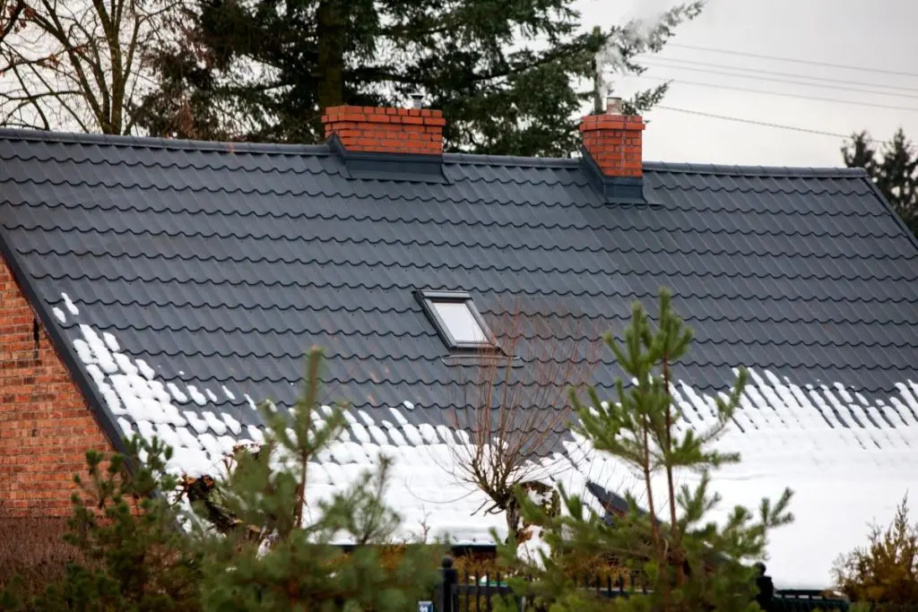 snow guards for metal roof  A sloped roof with dark gray tiles has two red brick chimneys and a skylight. Patches of snow cover the lower part of the roof. Evergreen trees partially obscure the view.