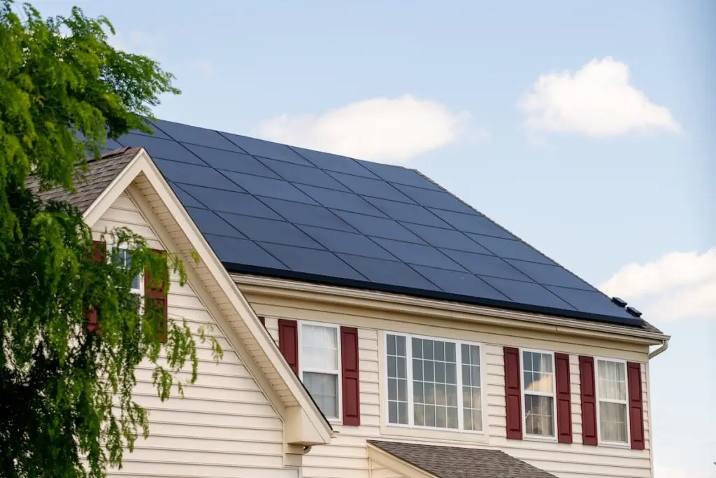 A charming two-story house in waunakee boasts white siding and red shutters, topped with a solar installation on the roof. The sky is clear with a few clouds, and a tree with lush green leaves is partially visible on the left.