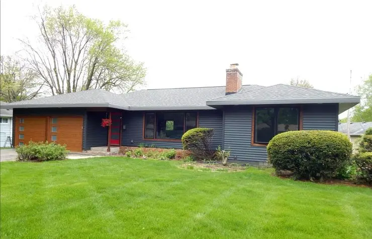 A modern single-story house with dark blue siding and a contrasting wooden garage door. The front yard features a neatly maintained lawn, landscaped shrubs, and a few trees in the background. Large windows and a red door add distinctive accents.