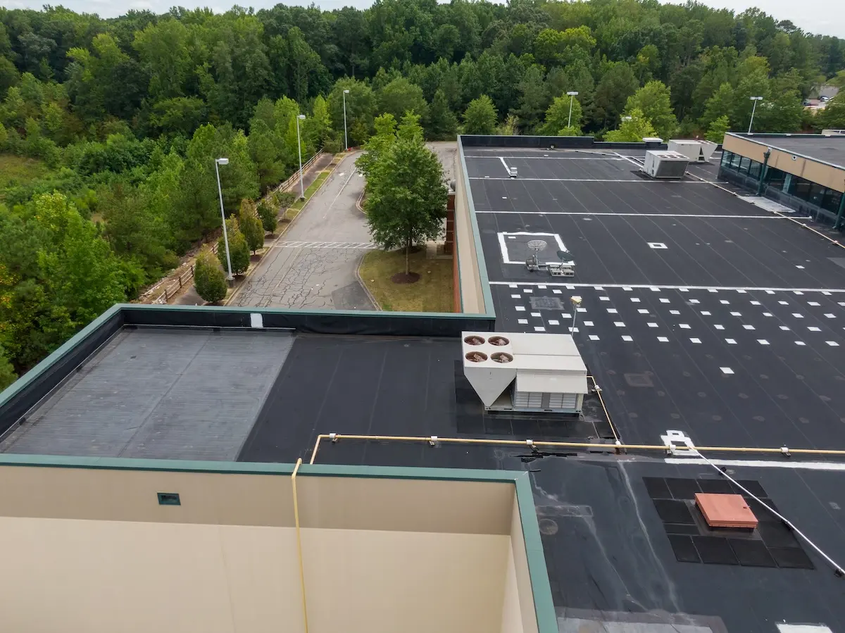 Aerial view of a vacant parking lot next to a large flat-roofed commercial building with rubber roofing, surrounded by dense green trees. The roof is equipped with HVAC units and other equipment.