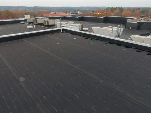 types of commercial roofing systems Flat black commercial rooftop with HVAC units and vents, surrounded by low parapet walls. Trees with autumn foliage are in the background under an overcast sky.