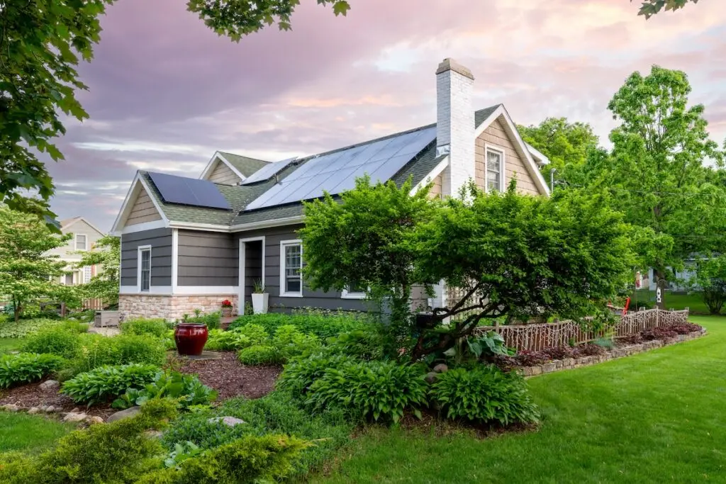 A charming house with gray siding and white trim is surrounded by lush greenery and garden plants. Solar panels are mounted on the roof. The sky is colorful with pink and purple hues, suggesting either sunrise or sunset.