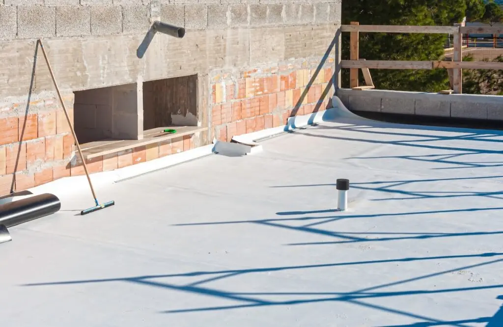 A flat rooftop under construction with a roofing membrane partially installed, tools and pipes scattered around, and unfinished brick and concrete walls enclosing the roof area. Professional workers waterproofing house construction site. Worker rolling membrane