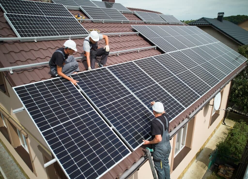 Three workers wearing safety gear skillfully install solar panels on the sloped roof of a suburban house in Middleton. Positioned strategically across the roof, they ensure each panel is securely placed, blending innovation with eco-friendly surroundings.