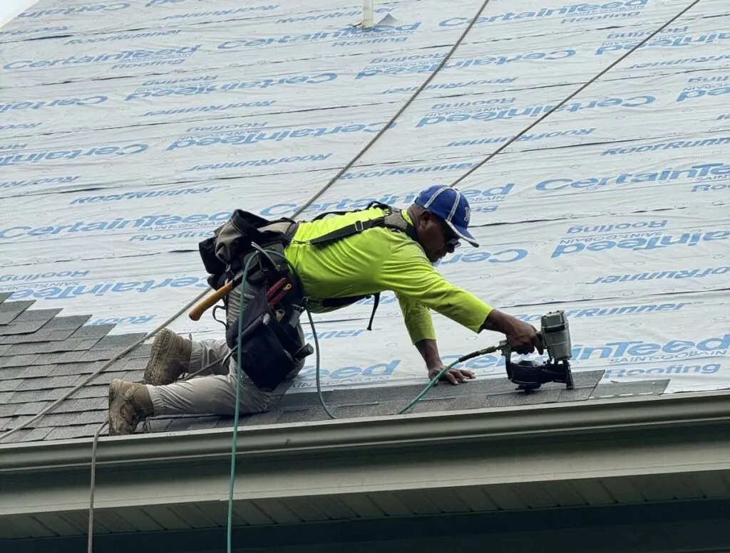 A worker in a neon green shirt and blue cap is kneeling on a roof, using a nail gun to secure shingles. He carries tools on his belt, with roofing materials and safety ropes visible around him.