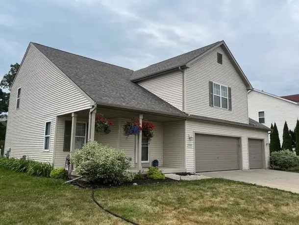 A modern, two-story house with beige siding, dark roof, and a two-car garage. The front porch features hanging flower baskets. The yard is landscaped with shrubs and a well-maintained lawn. The sky is overcast.