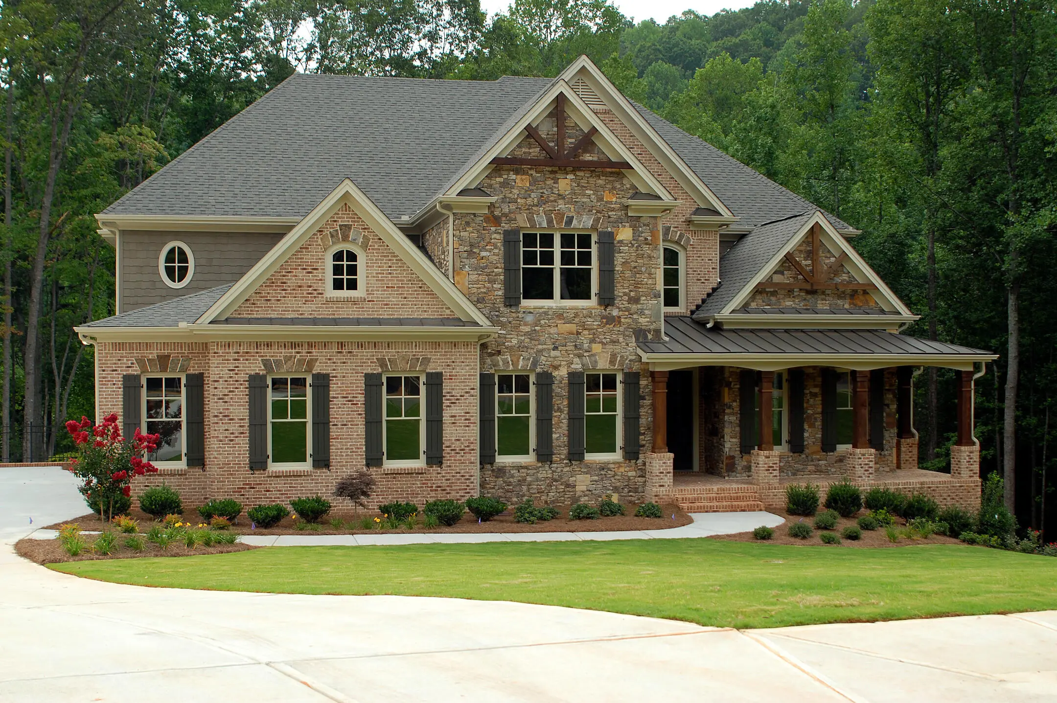 A large, elegant house with a mix of brick and stone facade, surrounded by lush greenery. It features a dark shingled roof, numerous windows, and a spacious front porch. The driveway leads up to the house through a neatly manicured lawn.