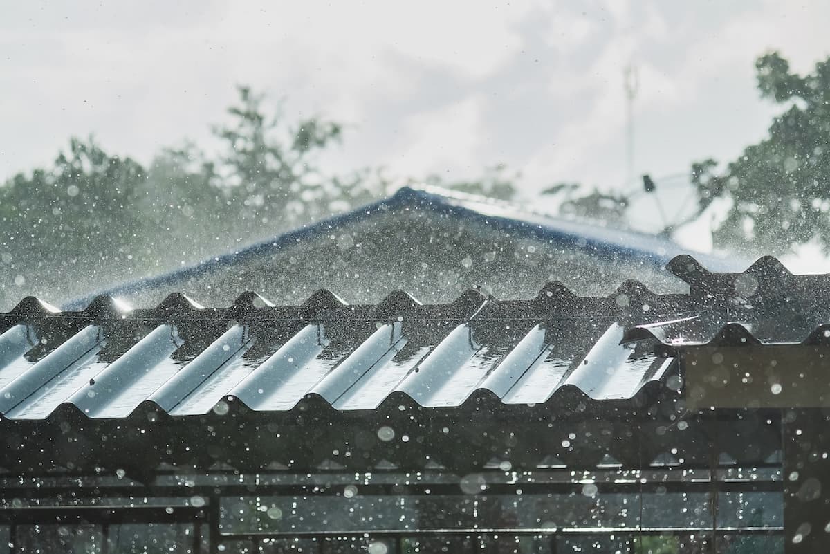 Raindrops splash and glisten on a corrugated metal roof, with blurred green trees and a house in the background, creating a fresh, rainy atmosphere.