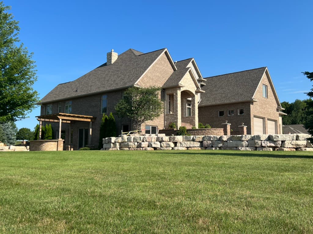 Two-story brick house with a large lawn, attached three-car garage, stone retaining wall, and a wooden pergola—showcasing expert roofing Fitchburg craftsmanship—set under a clear blue sky on a sunny day.