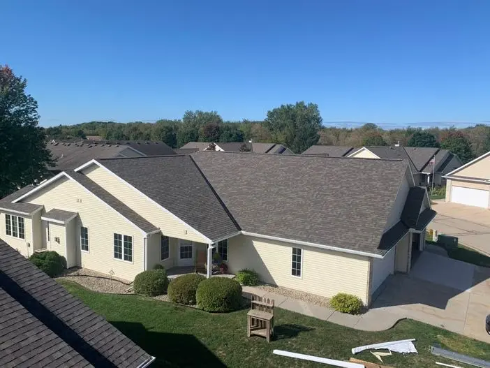 A row of single-story beige houses with dark gray roofs set against a backdrop of trees and a clear blue sky. The houses have neatly trimmed bushes and a driveway visible.