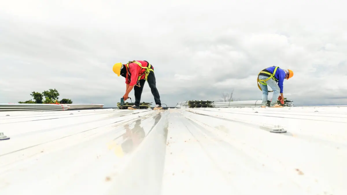 commercial roof repair services Two construction workers wearing safety gear and harnesses are working on a white metal roof under a cloudy sky. One is crouched, using tools, while the other is bent over, both focused on their tasks.