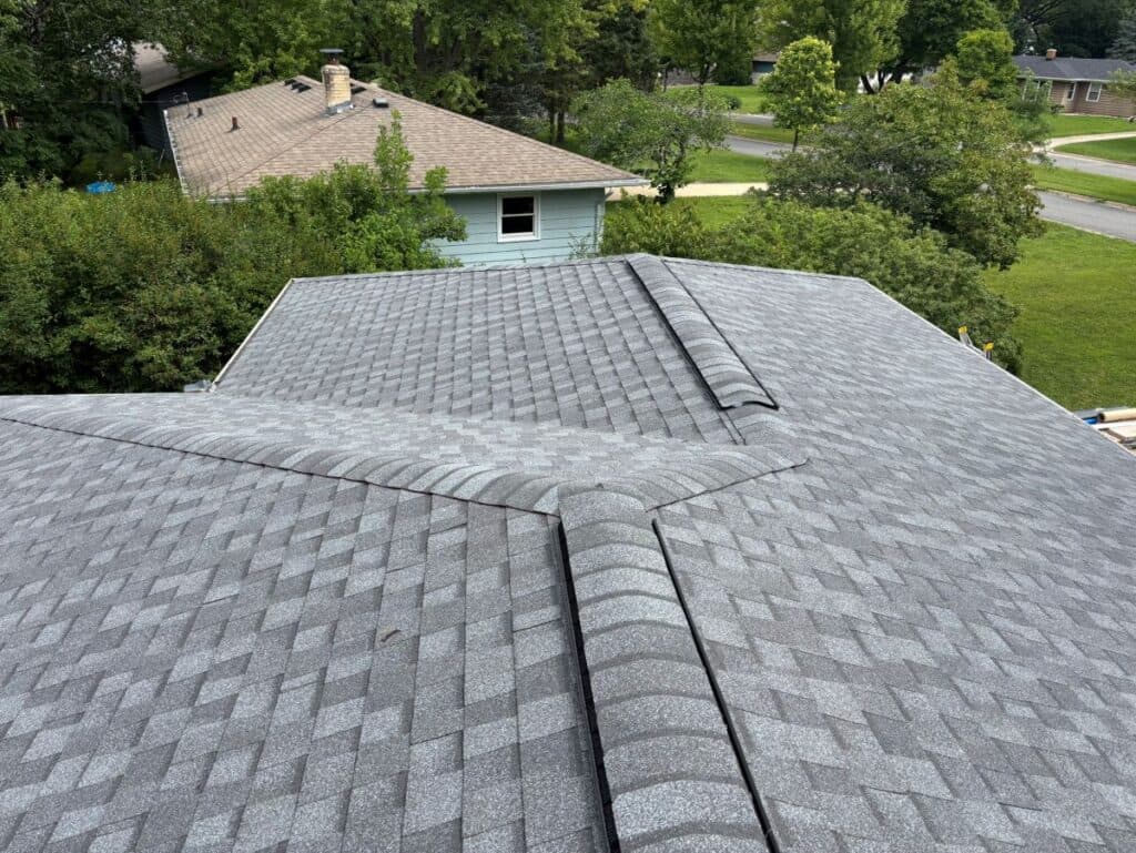 Aerial view of a house roof covered with gray asphalt shingles by roofing Waunakee experts, featuring intersecting ridges and surrounded by trees and neighboring houses in a suburban area.