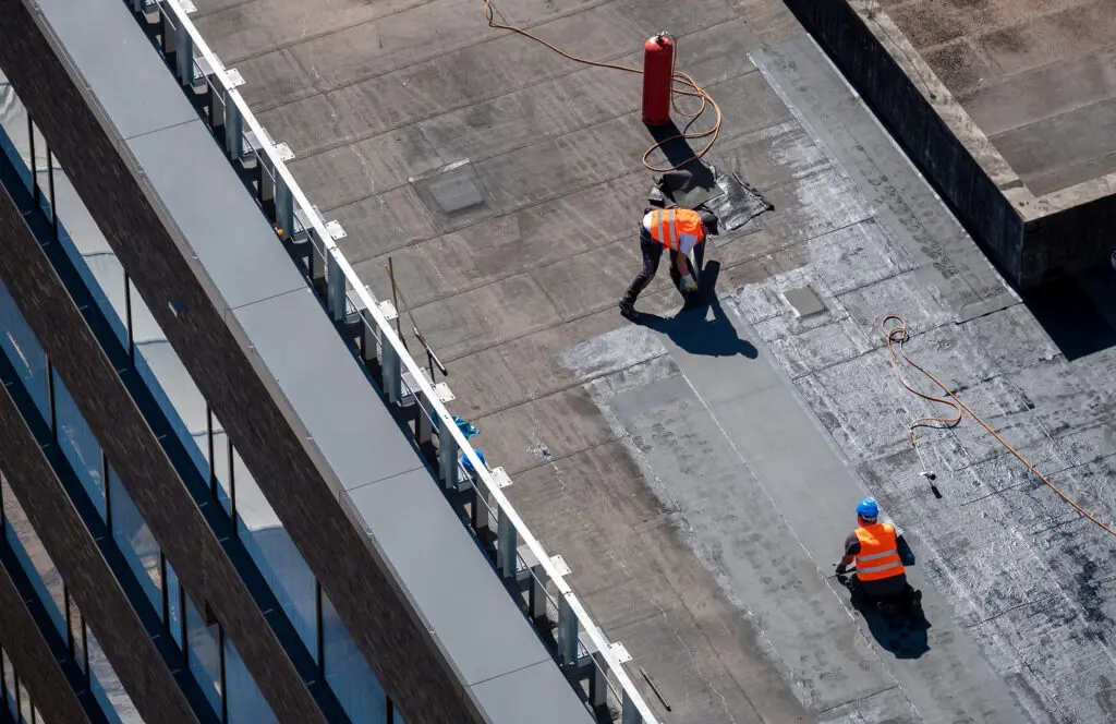 commercial flat roof repair Aerial view of three construction workers in orange vests and helmets working on a flat rooftop, with tools and equipment visible around them.