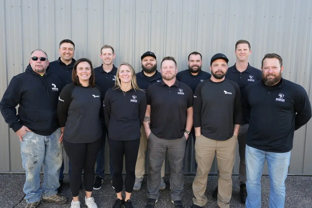 roofing companies sun prairie wi Twelve people in black shirts and jackets pose together in front of a corrugated metal wall, smiling at the camera.