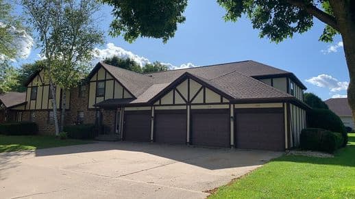 A large house with a Tudor-style facade featuring brown wood accents and cream panels. It has three garage doors, a gabled roof, and is surrounded by trees and greenery. The sky is clear with a few clouds.
