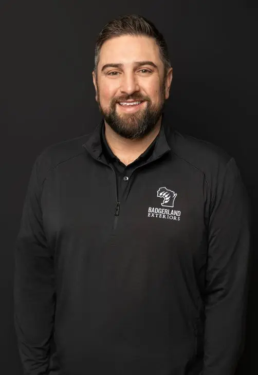 A man with short brown hair and a beard smiles while wearing a black quarter-zip pullover with the 