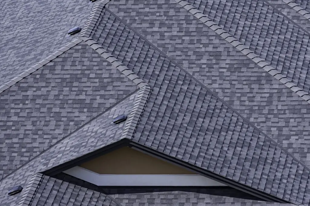A close-up view of overlapping grey architectural shingles on rooftops, featuring repeating geometric patterns and textures with angled surfaces and clean lines.